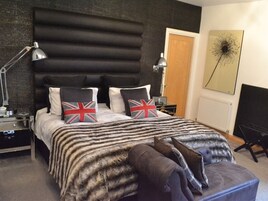 Quarto com cama grande, cabeceira, duas luminárias de cabeceira, mesa de apoio, uma cadeira e uma obra de arte na parede.