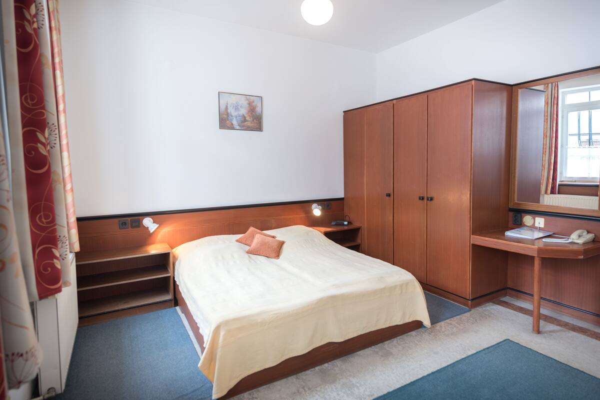 A hotel room with a bed, a desk, a wardrobe, a painting on the wall, and a window with curtains.