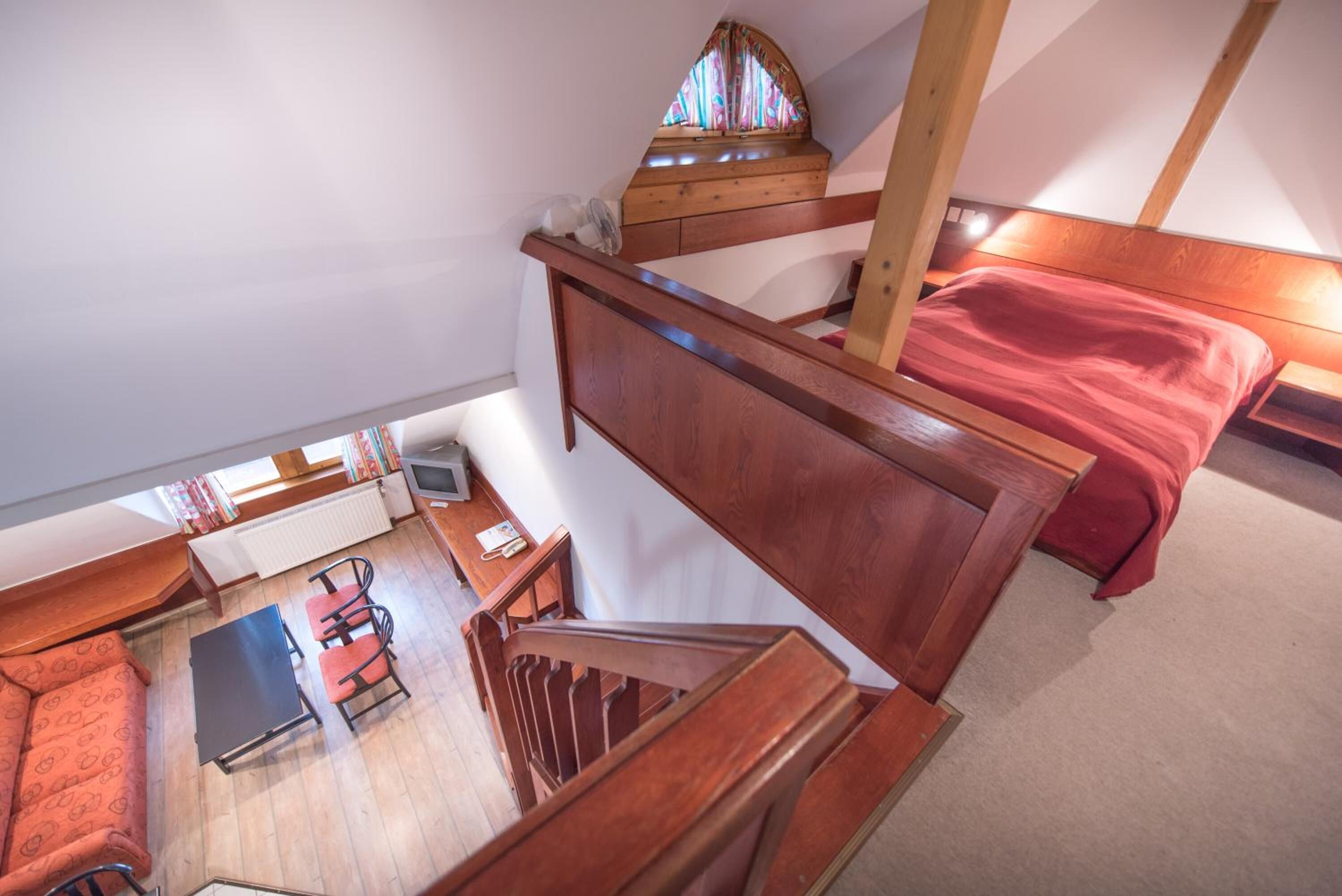 A loft-style bedroom with a wooden staircase, a bed with a red blanket, a small desk with a chair, and a window with a colorful view.
