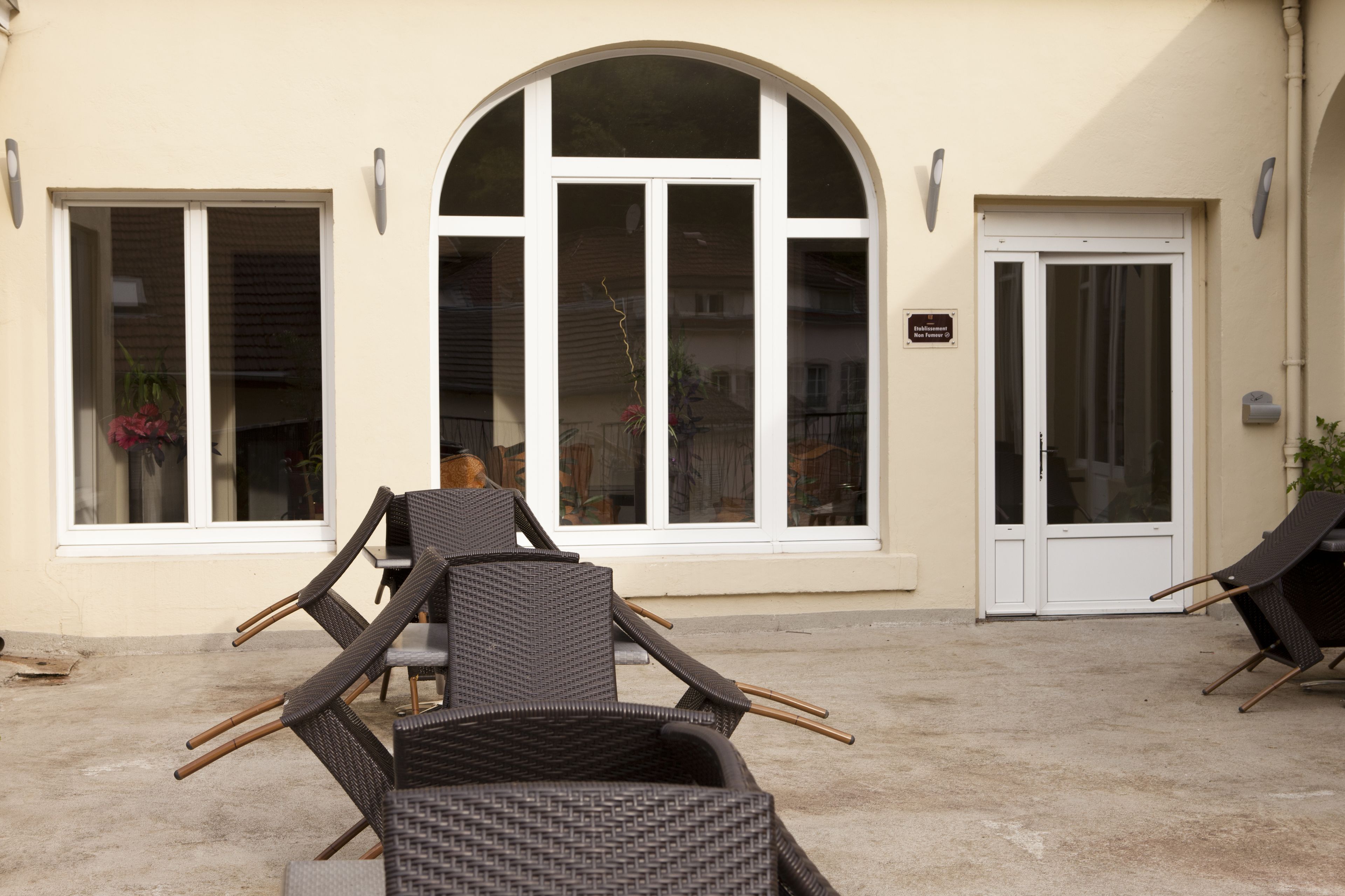 An outdoor seating area with wicker chairs and a table, a large arched window, and a door with a sign.