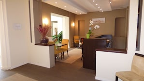 A hotel lobby with a reception desk, a dining area with chairs and a table, and a hallway leading to the bathroom. - Le Beausejour (Plombieres-les-Bains)