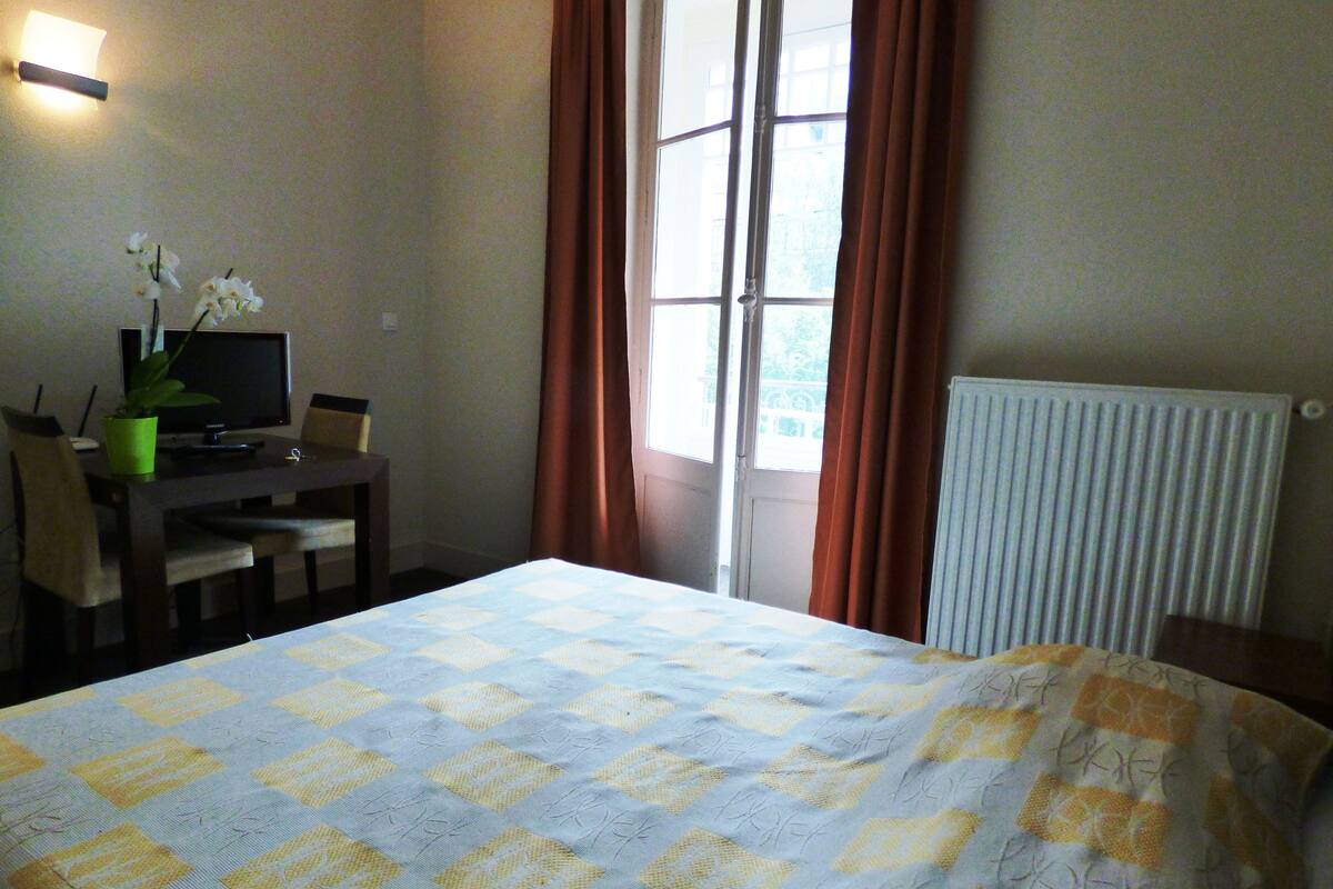 Habitación de hotel con una cama, un escritorio con computadora, una silla, una planta en maceta y una ventana con cortinas.