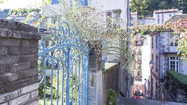A narrow street with a blue wrought iron gate, stone walls, and buildings with balconies. - Le Beausejour (Plombieres-les-Bains)