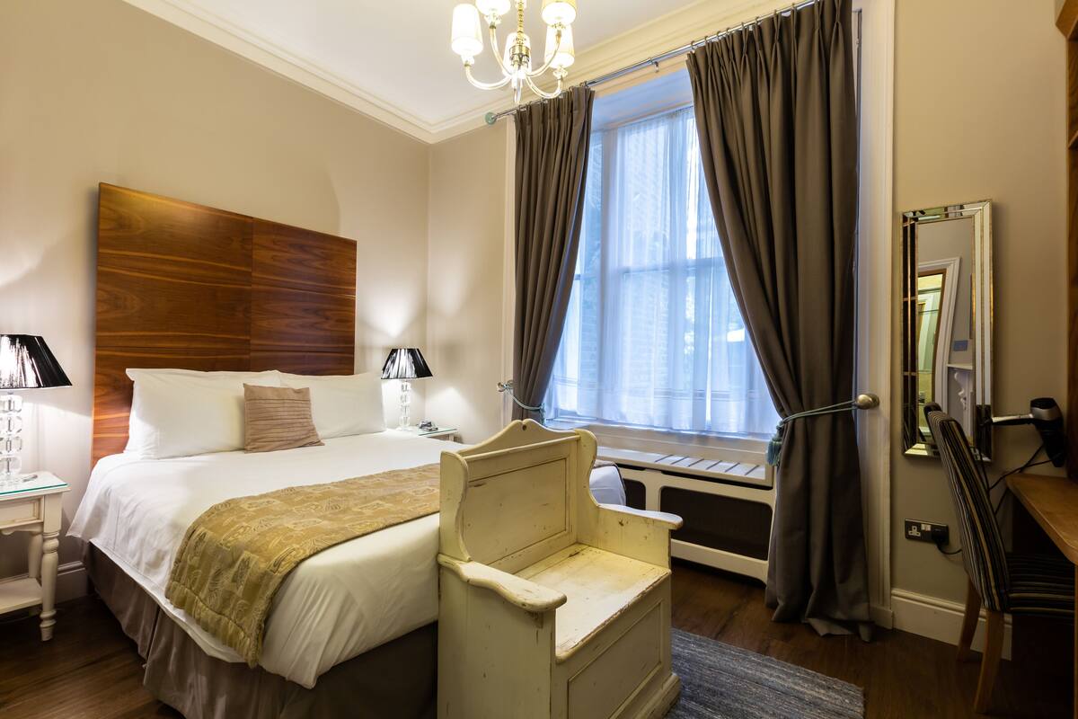 A hotel room with a large bed, a bedside table with a lamp, a chair, a window with curtains, and a mirror.
