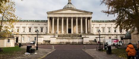 A grand neoclassical building with a central dome and columns, a paved pathway leading to the entrance, and a few people in the foreground.