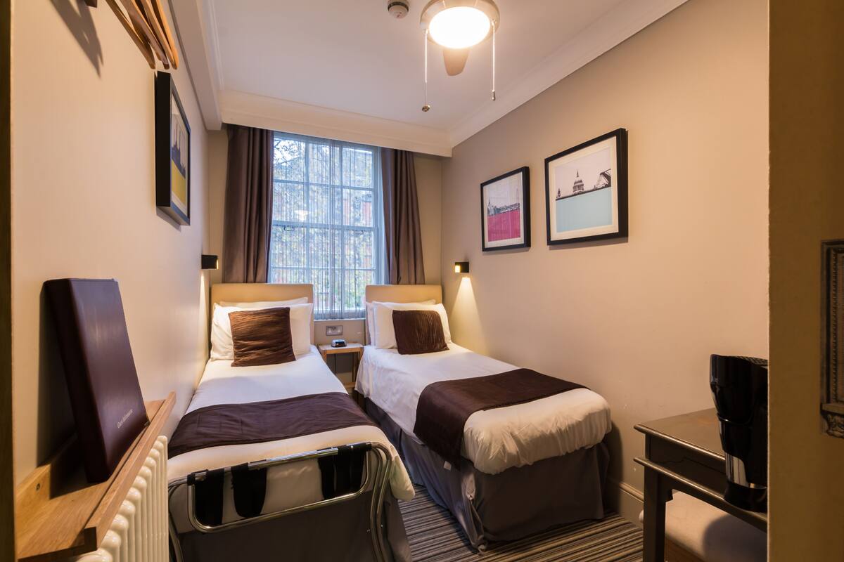 A hotel room with two beds, a desk, a chair, a radiator, and a window with curtains.