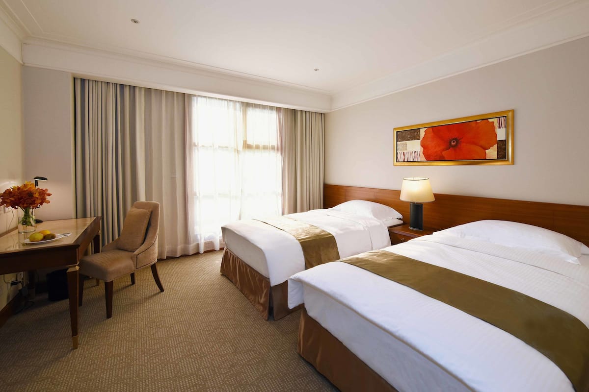 Quarto de hotel com duas camas, uma escrivaninha com cadeira, uma luminária e uma janela grande com cortinas.