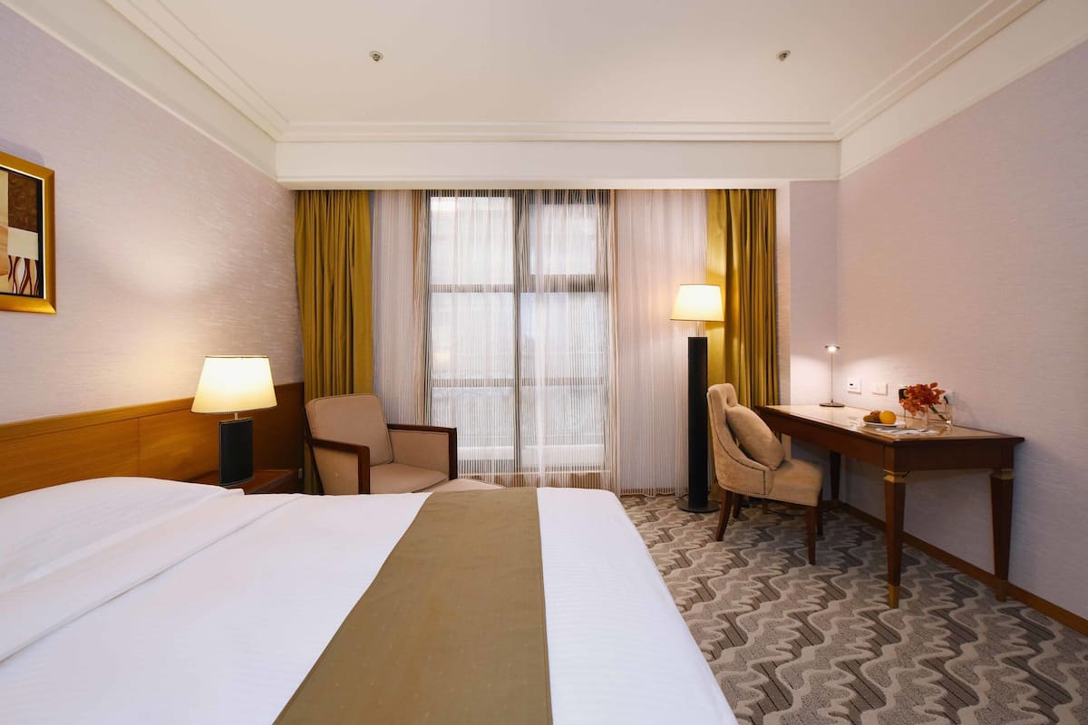 Quarto de hotel com uma cama grande, duas cadeiras, uma mesa de escritório e uma janela com cortinas.