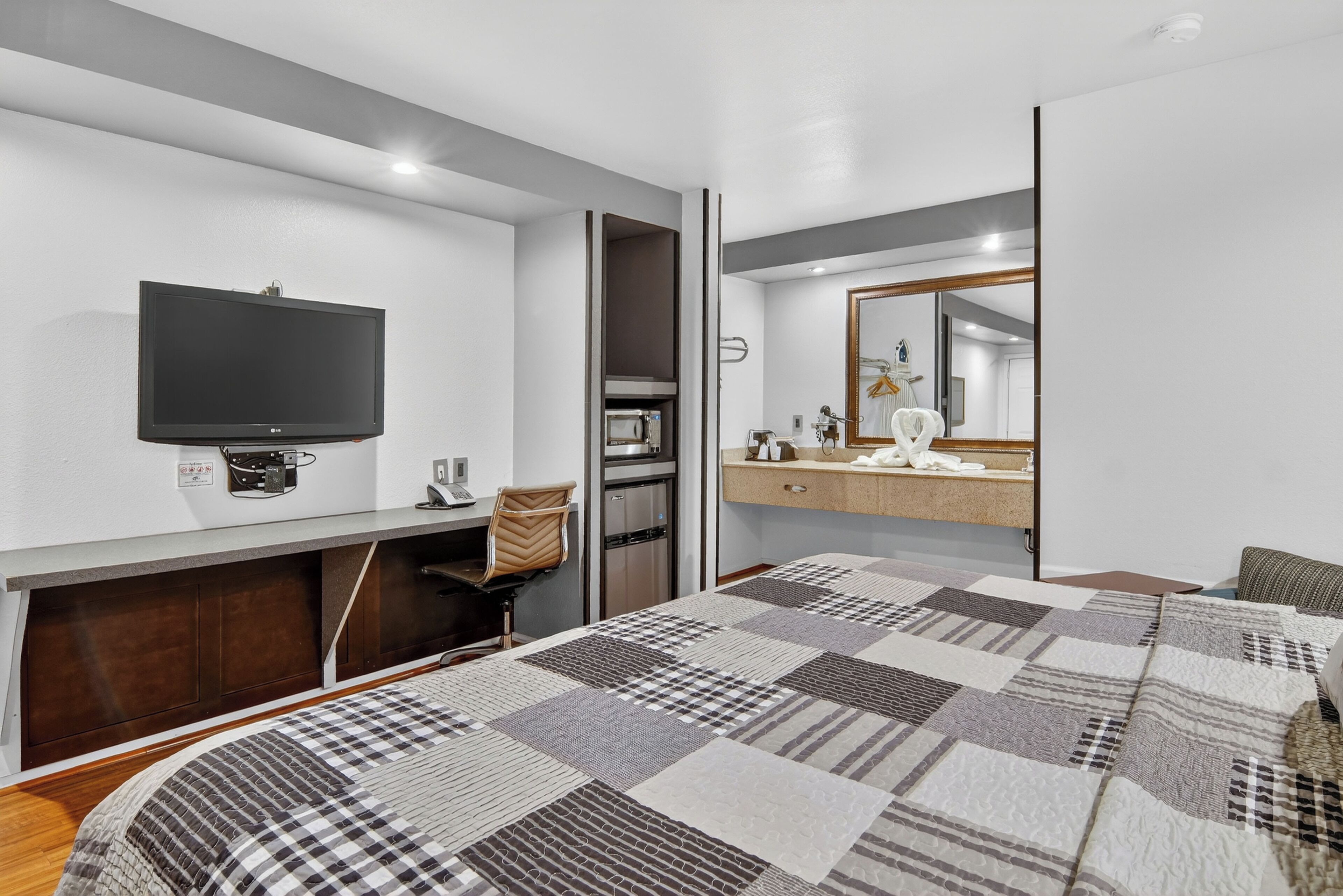 A modern hotel room with a large bed, a flat-screen TV, a microwave, a coffee machine, and a bathroom with a mirror and towel rack.