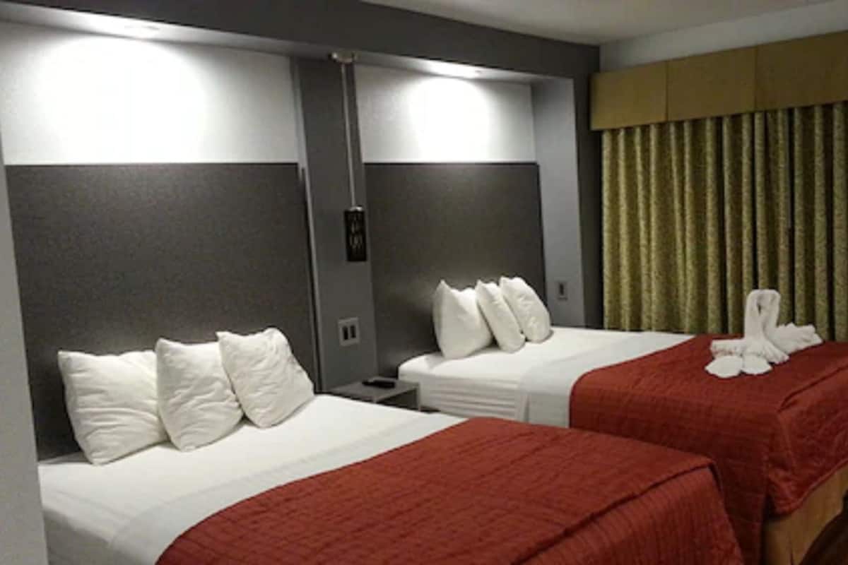 A hotel room with two beds, each with white bedding and red blankets. There are white pillows on each bed. The room has a neutral color scheme with grey walls and a brown headboard. A curtain is visible on the right side of the image.