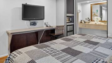 Une chambre d’hôtel comprenant un lit, un bureau avec un téléviseur à écran plat, une chaise, un minibar et une salle de bain avec un miroir et un porte-serviettes.