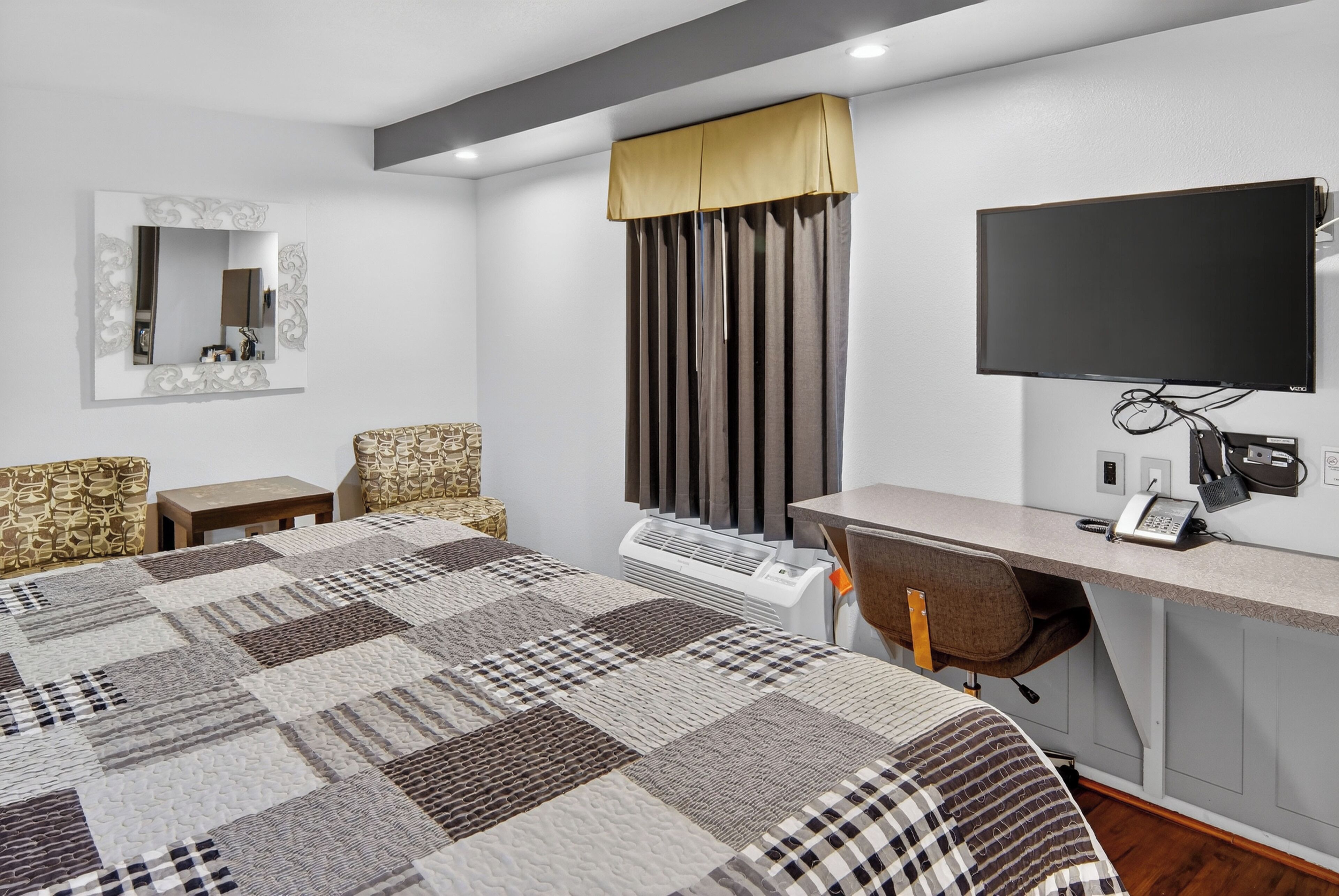 A hotel room with a large bed, a desk with a chair, a flat-screen TV, a mirror, and a window with curtains.