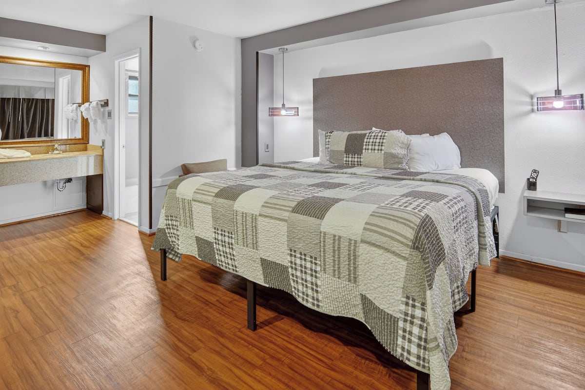 A modern bedroom with a large bed, a wooden floor, a bathroom with a mirror and sink, and a bedside table with a lamp.