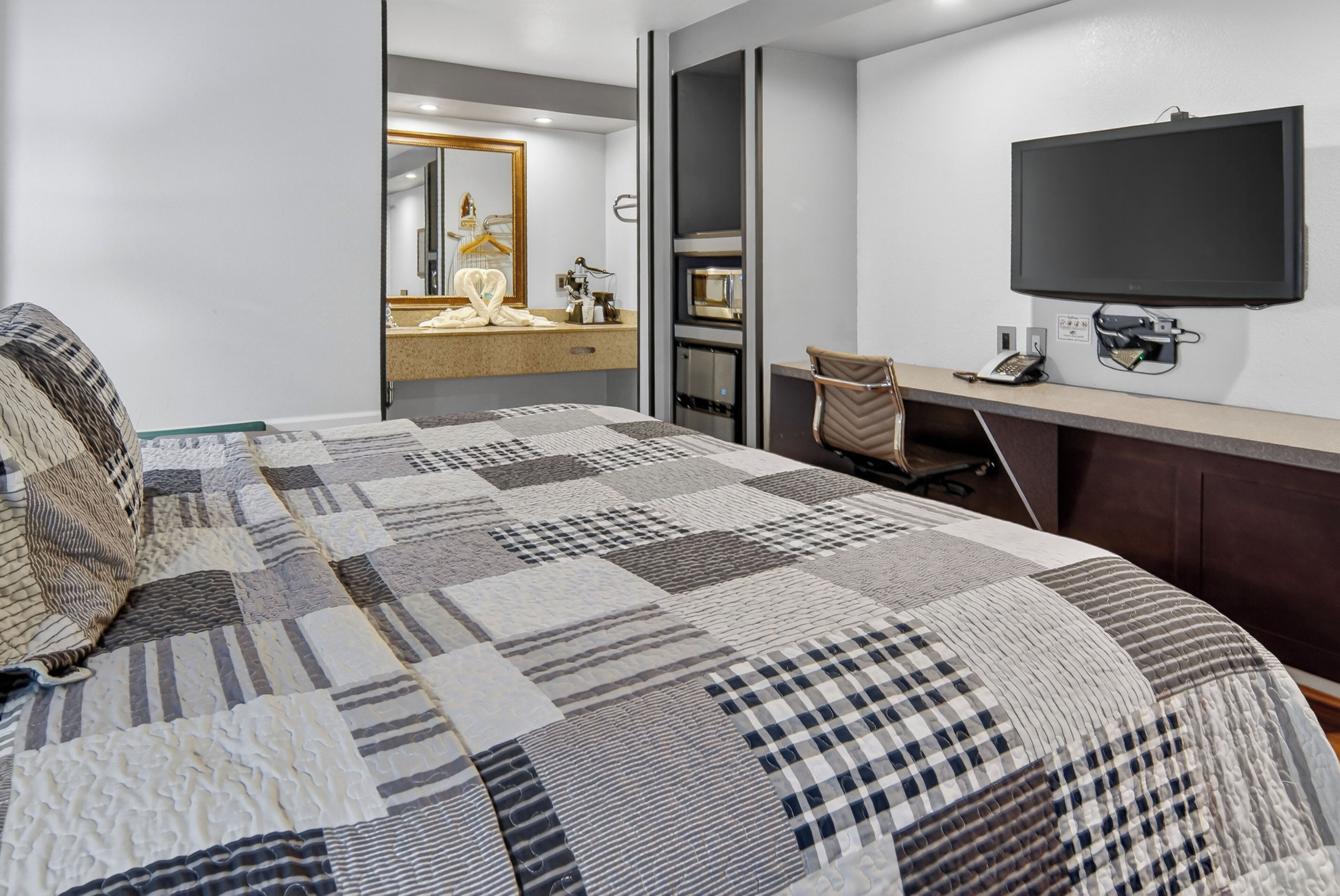 A modern hotel room with a large bed, a flat-screen TV mounted on the wall, a desk with a telephone, and a mirror above the vanity.