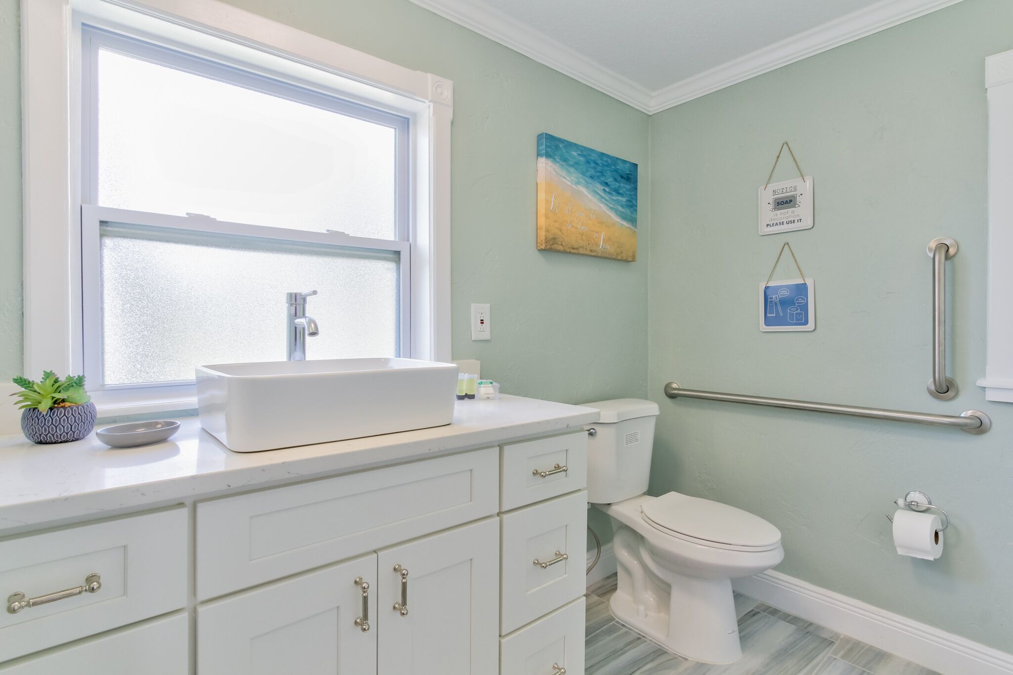 Une salle de bain avec un lavabo blanc, des toilettes, une fenêtre et une barre d’appui.