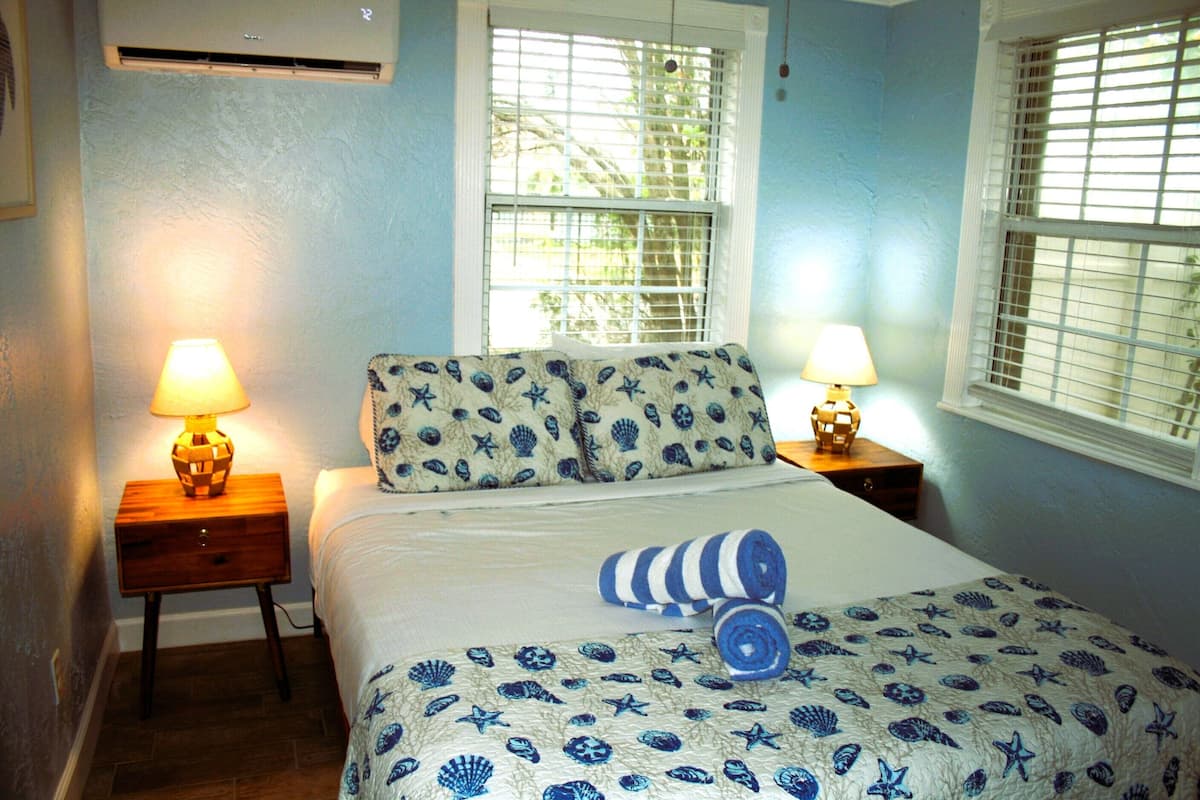 A bedroom with a bed, two nightstands with lamps, a window with blinds, and a patterned bedspread.