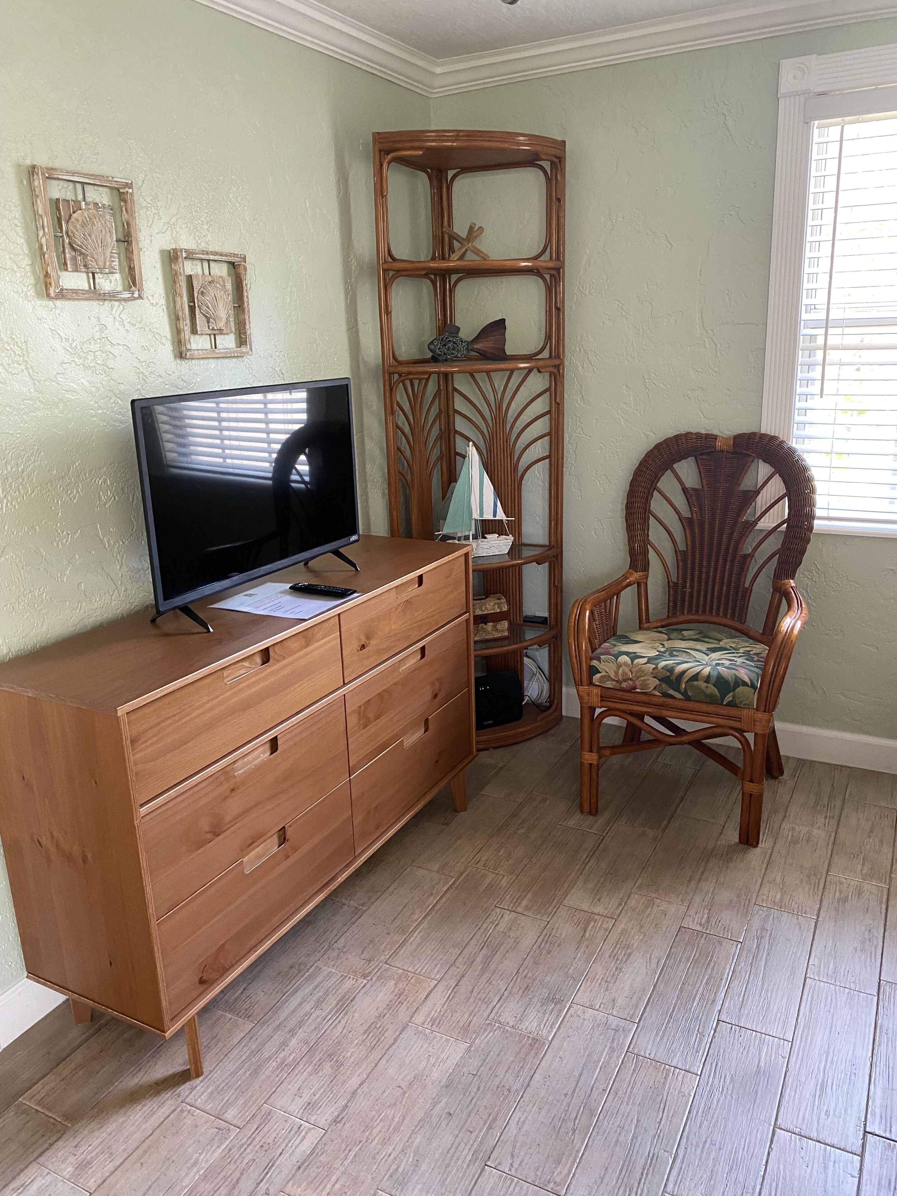 A room with a wooden dresser, a television on top, a wicker chair with a floral cushion, and a decorative wooden shelf with a small sailboat model.