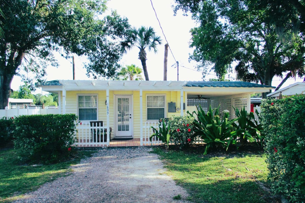 A yellow single-story house with a front porch, a gravel driveway, and surrounding greenery.