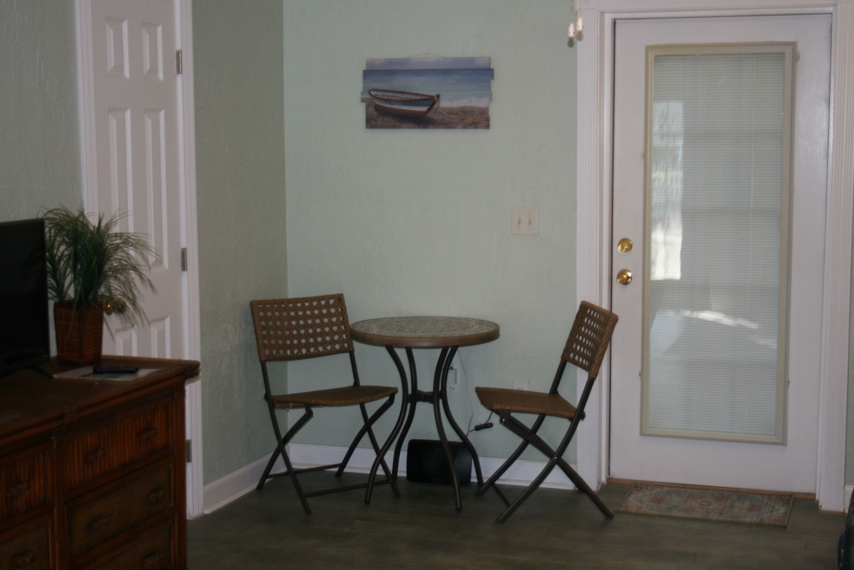 A room with a white door, a small round table, two wooden chairs, a television on a wooden cabinet, and a potted plant.