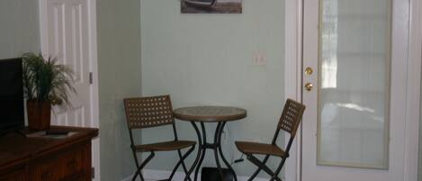 A room with a white door, a small round table, two wooden chairs, a television on a wooden cabinet, and a potted plant.
