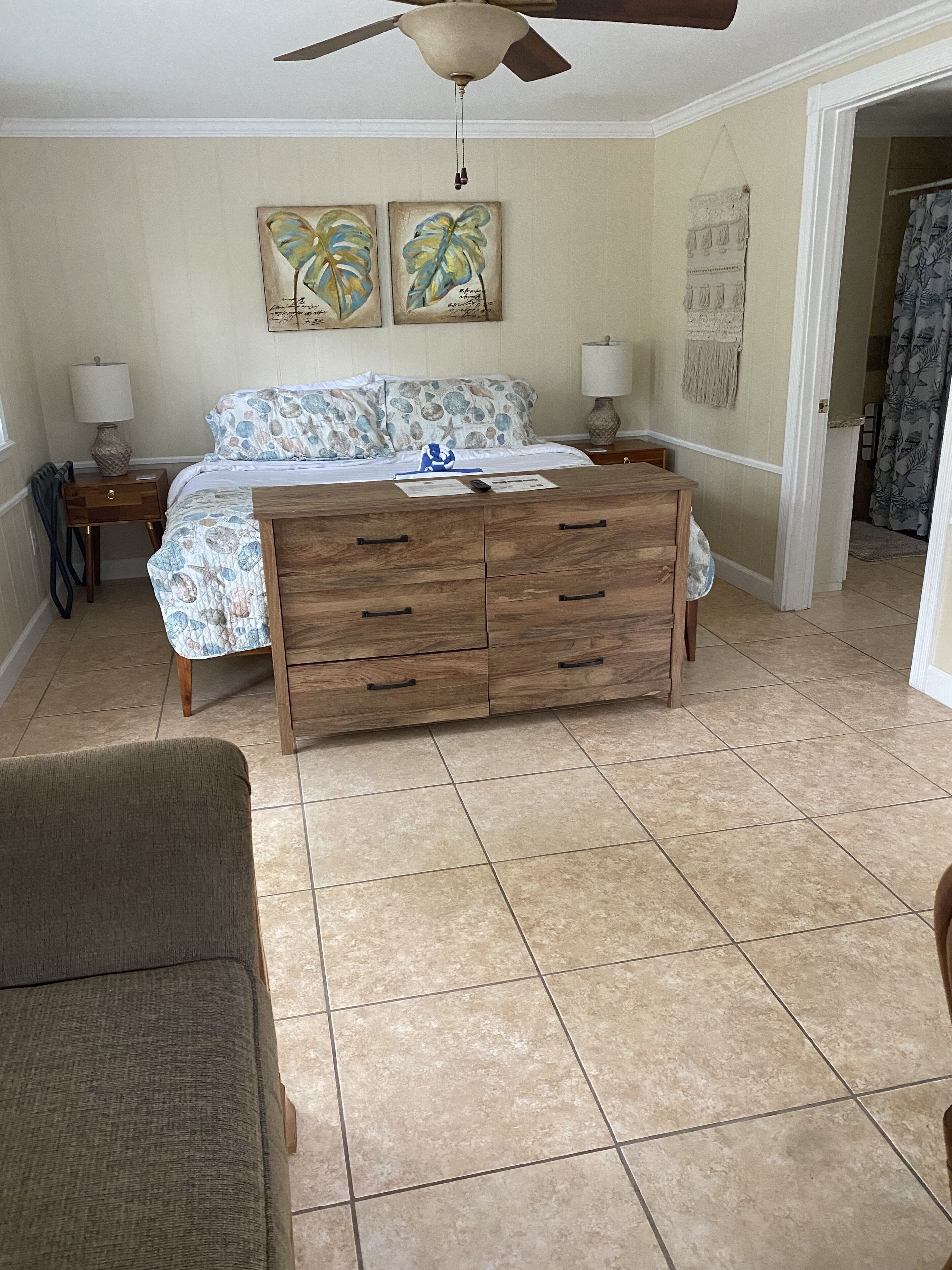 A bedroom with a bed, wooden dresser, two wall art pieces, and a ceiling fan.