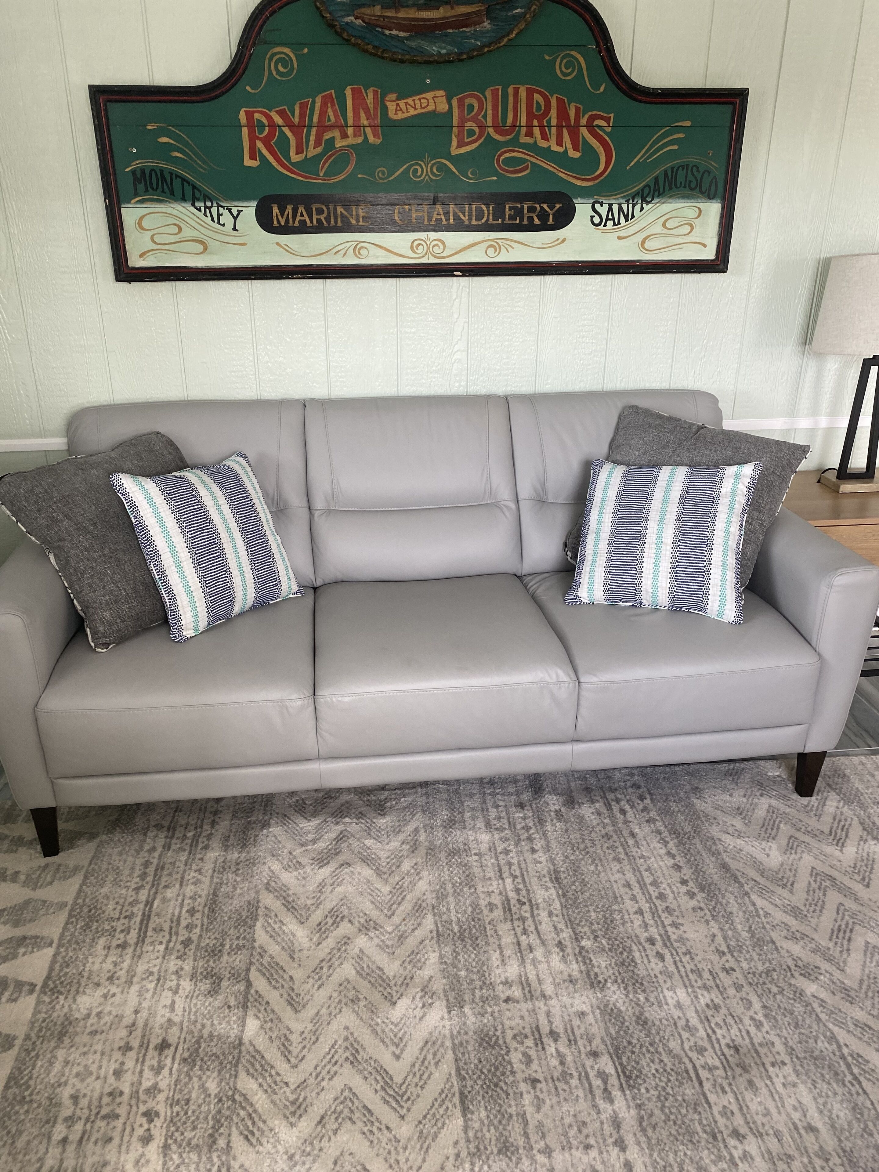 A grey leather sofa with patterned cushions against a herringbone-patterned rug. A vintage sign above reads "RYAN AND BURNS MARINE CHANDLERY SAN FRANCISCO".