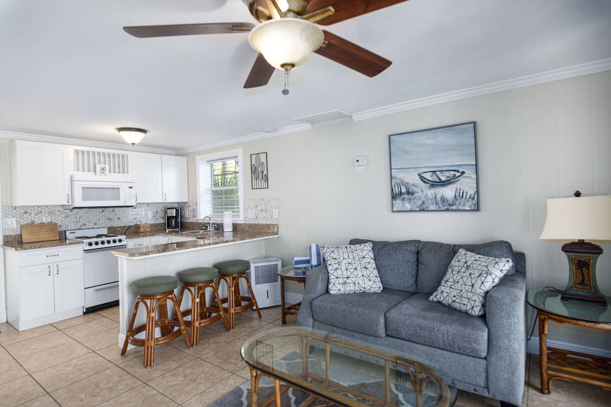 A modern kitchen with white cabinets, a microwave, and a stove. There is a gray sofa with patterned cushions and a glass coffee table. A ceiling fan is visible above the kitchen.