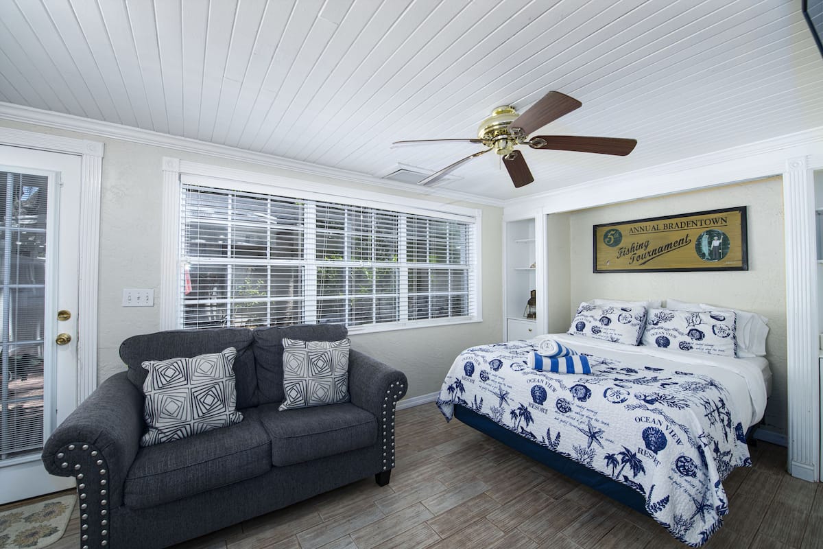 A bedroom with a bed, a gray sofa, a ceiling fan, and a window with blinds.