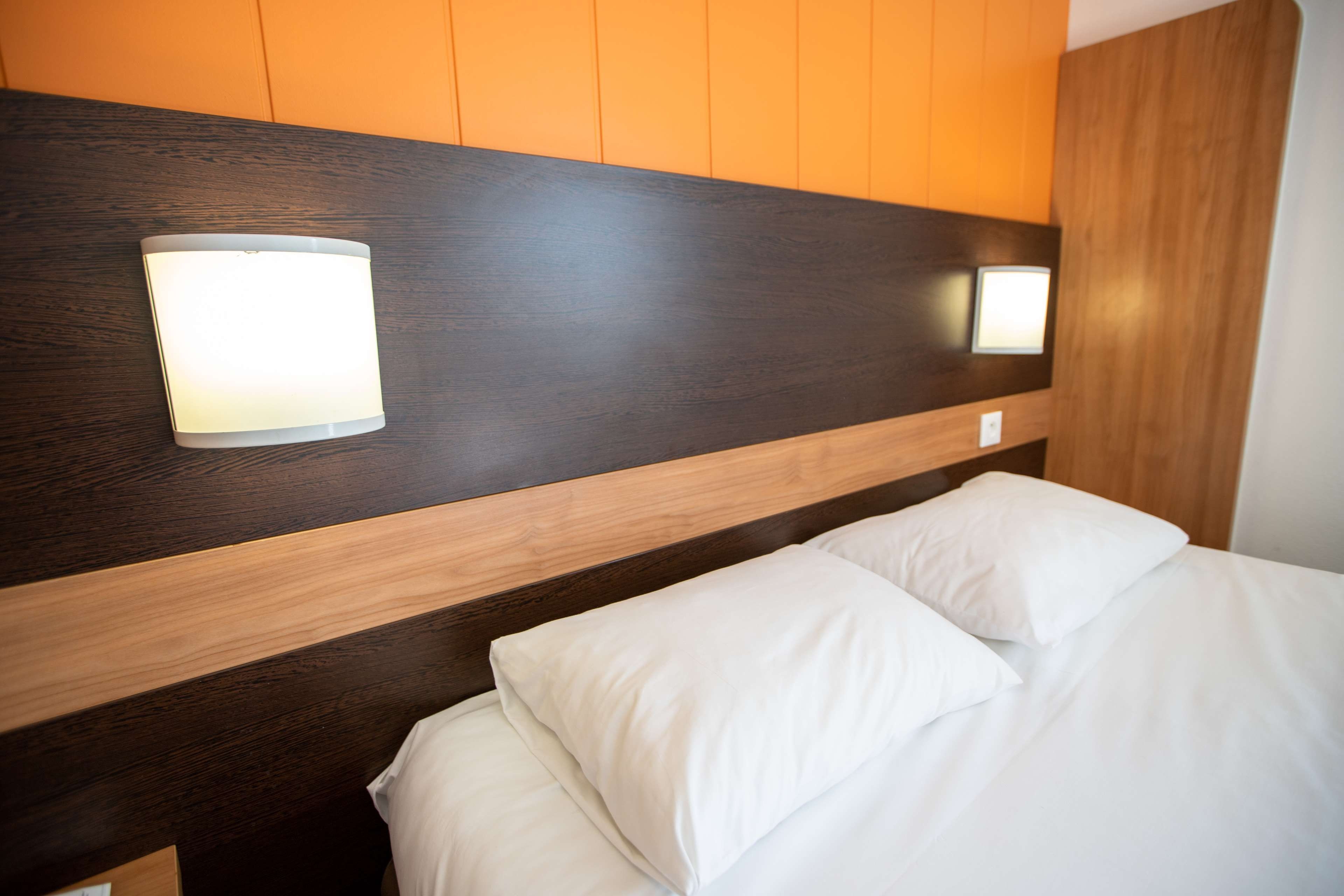 Une chambre d’hôtel avec une tête de lit en bois, deux lampes fixées au mur et un lit avec une literie blanche.