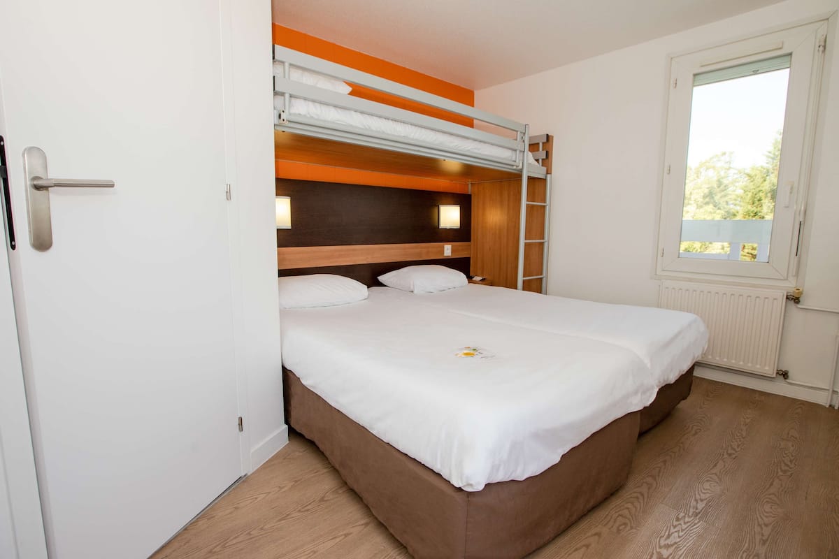 A single-bed room with a bunk bed above, wooden flooring, a white door, a window with a view of greenery, and two wall-mounted lights.