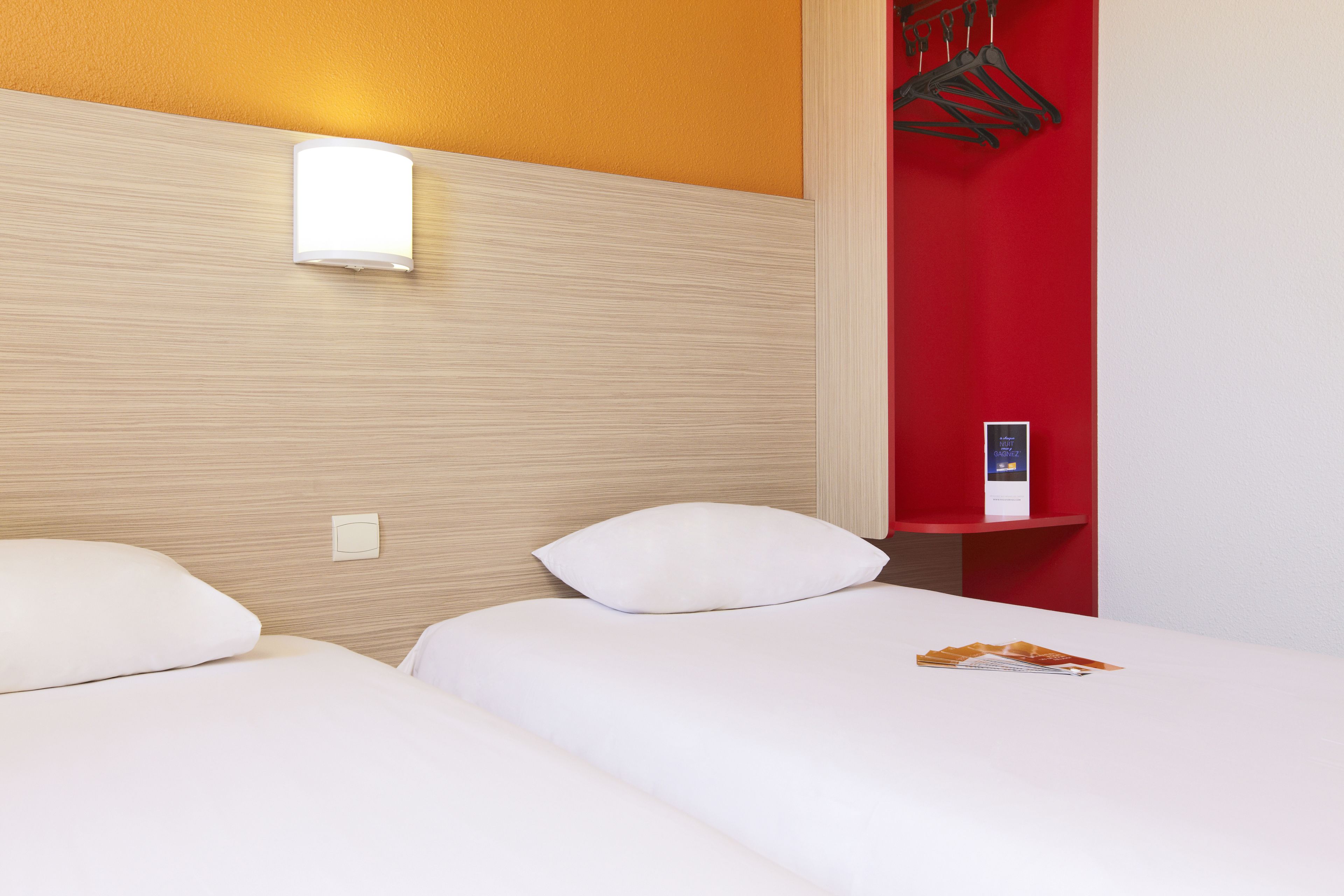 Une chambre d’hôtel avec deux lits, un mur d’accent rouge, une table de chevet avec une lampe et un porte-manteau.