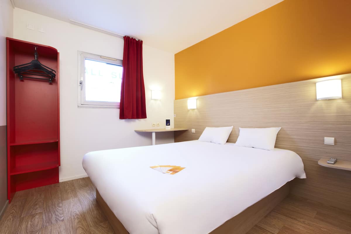 A hotel room with a large bed, a red wardrobe, a window with red curtains, and two wall-mounted lights.