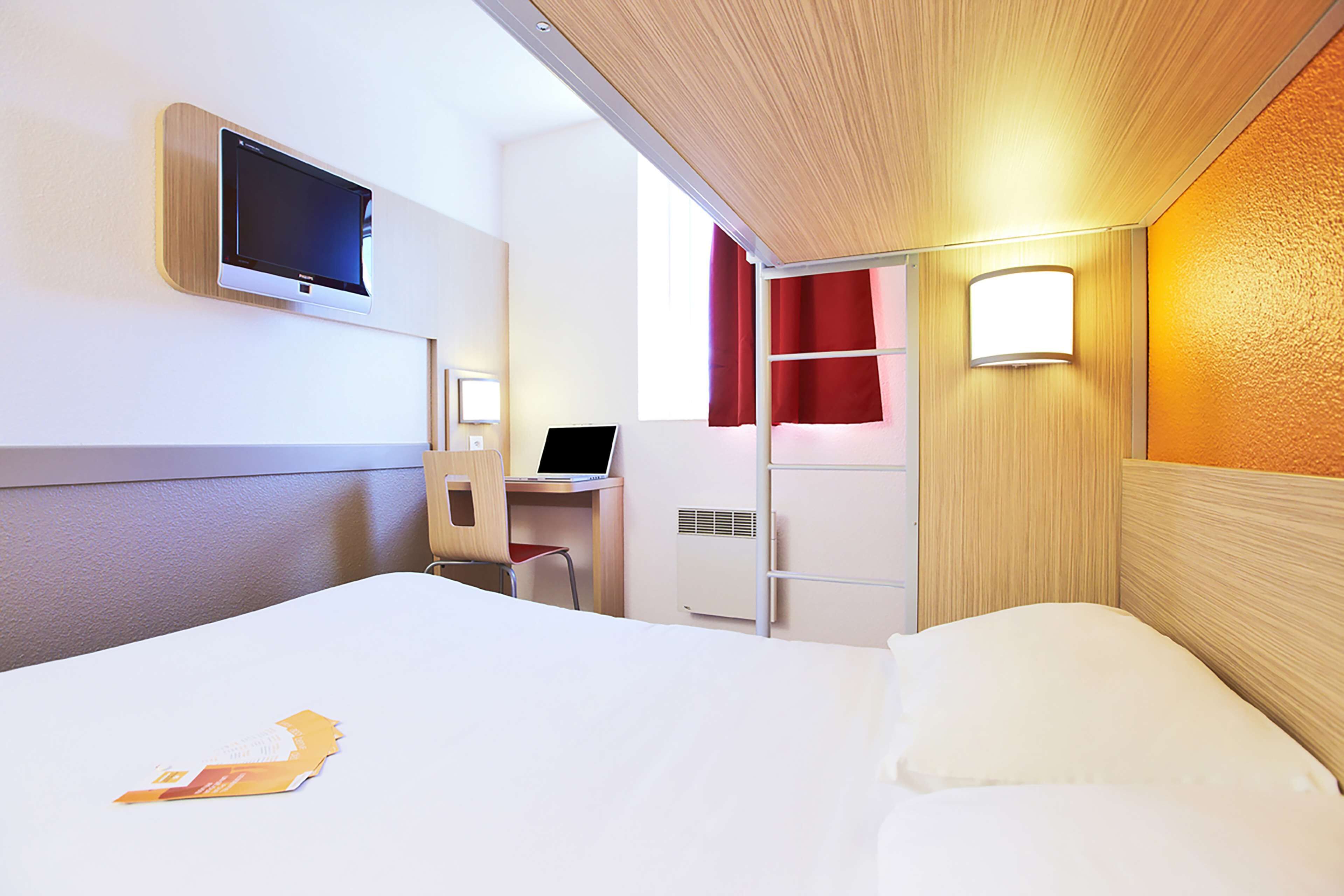 A hotel room with a bed, a desk with a laptop, a TV mounted on the wall, and a window with red curtains.