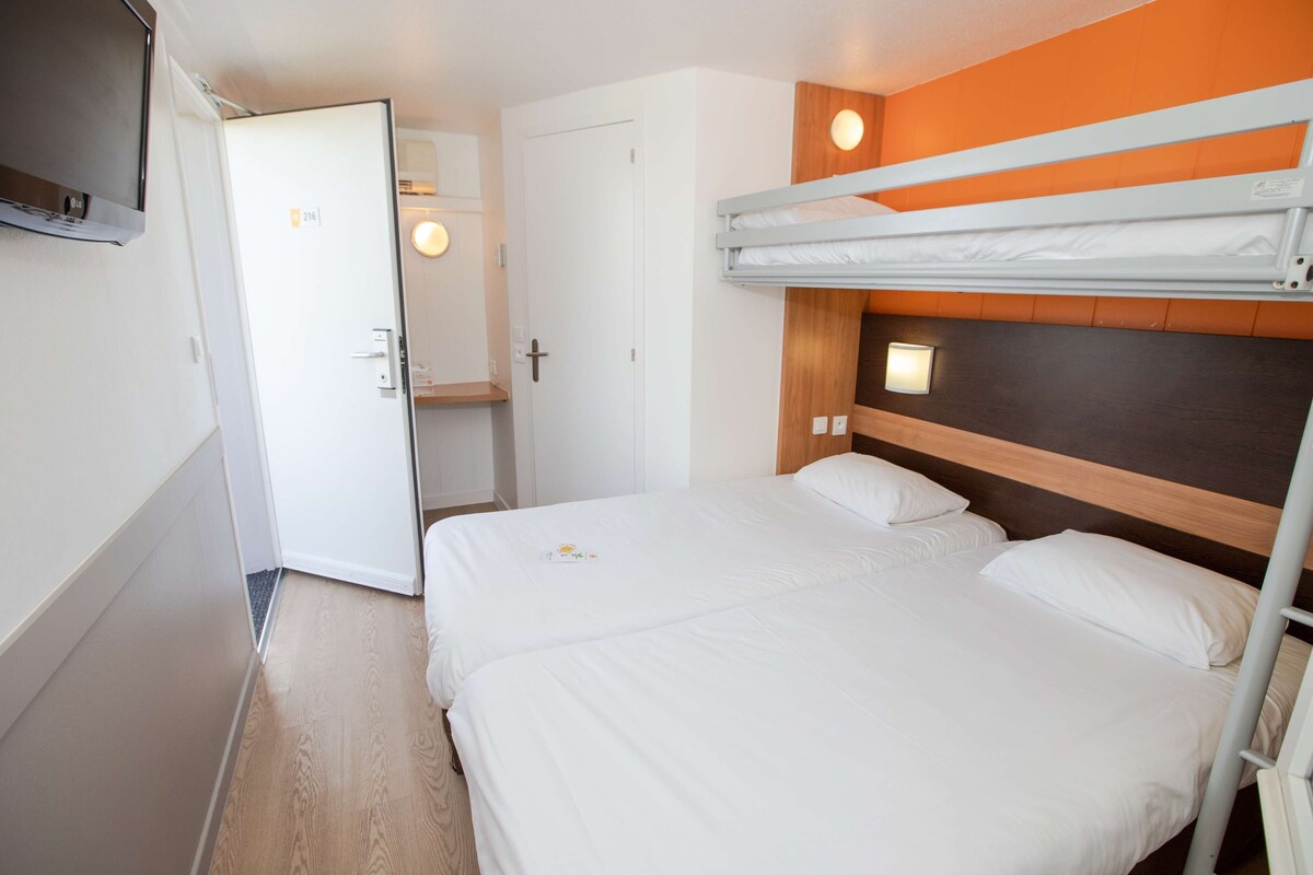 A hotel room with a bunk bed, a white door, a small bedside table, and a wall-mounted TV.