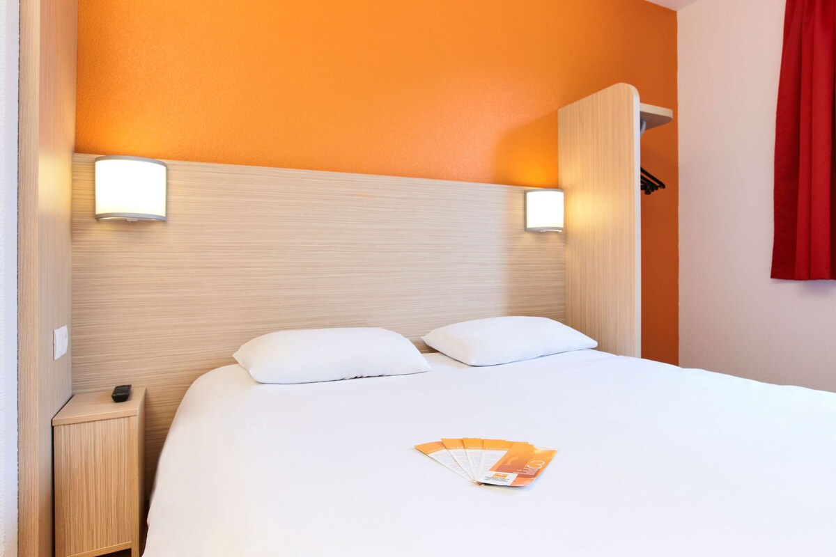 A hotel room with a bed, two pillows, a nightstand, a red curtain, and a wall-mounted light fixture.