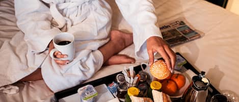 Une personne vêtue d’une robe, assise sur un lit, avec un plateau de nourriture devant elle, comprenant du pain et un muffin, ainsi qu’une tasse de café.