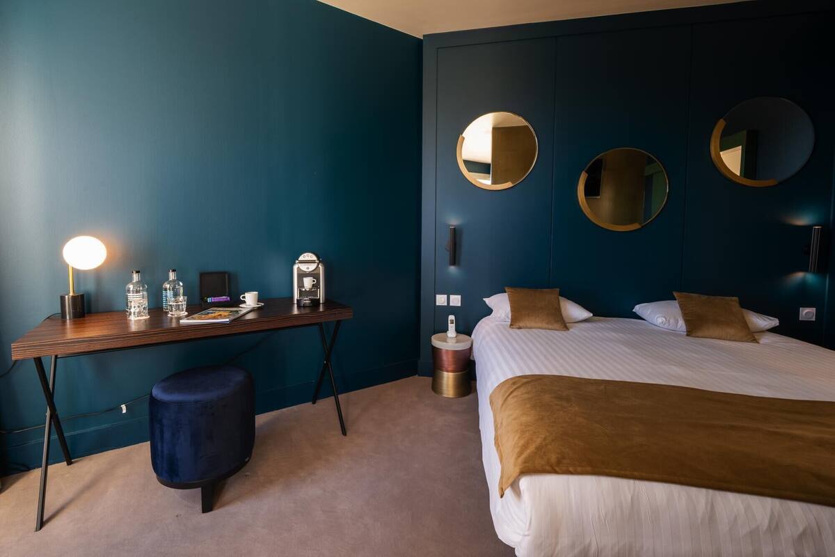 A hotel room with a bed, bedside tables, a desk with a coffee machine, and two wall-mounted mirrors.