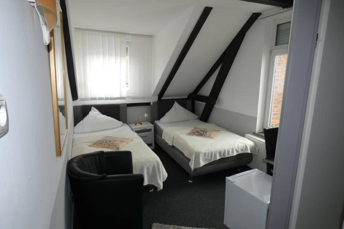 A compact bedroom with two single beds, a small bedside table with a lamp, a window with blinds, and a sloped ceiling with exposed beams.
