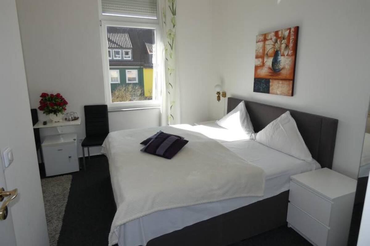 A hotel room with a bed, bedside tables, a desk with a chair, a window with curtains, and a painting on the wall.