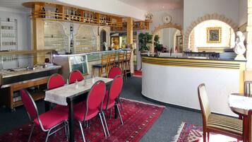 Un restaurante con zona de bar, sillas tapizadas en rojo y alfombra estampada.