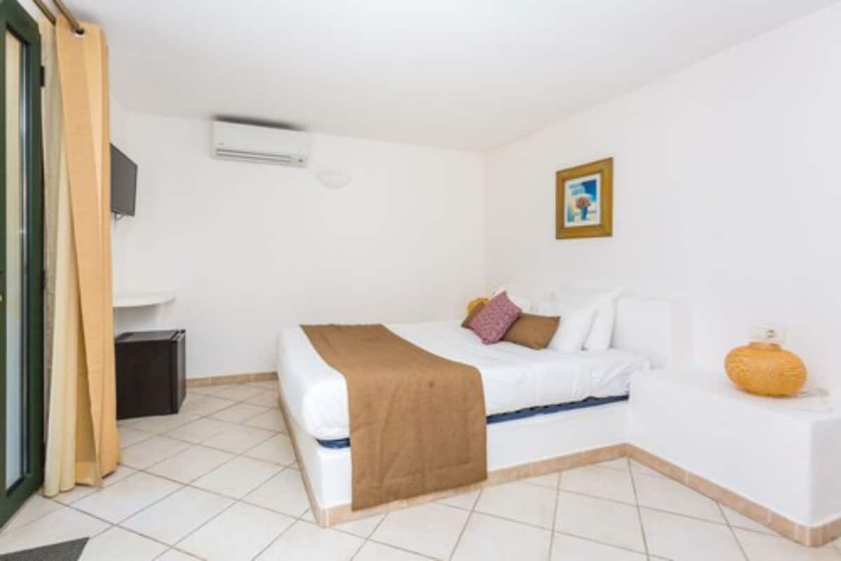 A hotel room with a bed, a bedside table, a small cabinet, a picture on the wall, and an air conditioning unit.