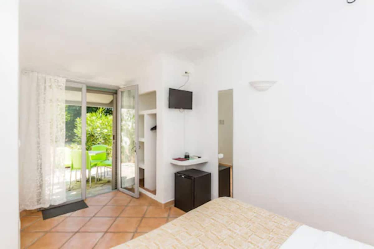 A bedroom with a bed, a small desk, a television, and a sliding glass door leading to an outdoor area with a chair and table.