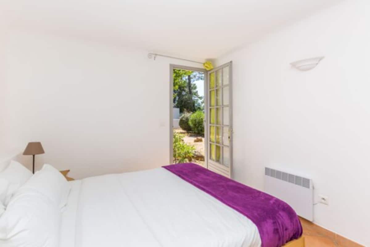 A bedroom with a bed, a purple throw, a window, and a view of greenery outside.