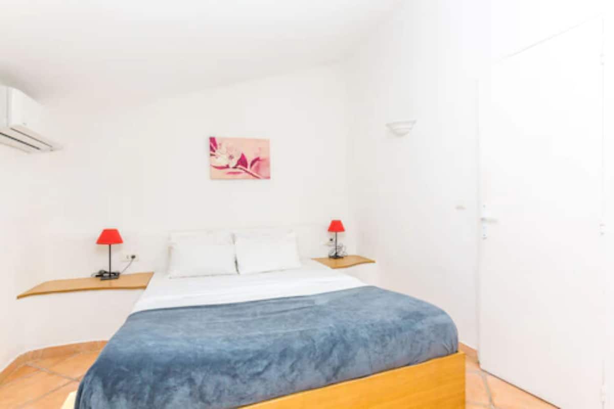 A bedroom with a bed, two bedside tables, a red lamp on each, a wall-mounted air conditioner, and a small pink artwork.