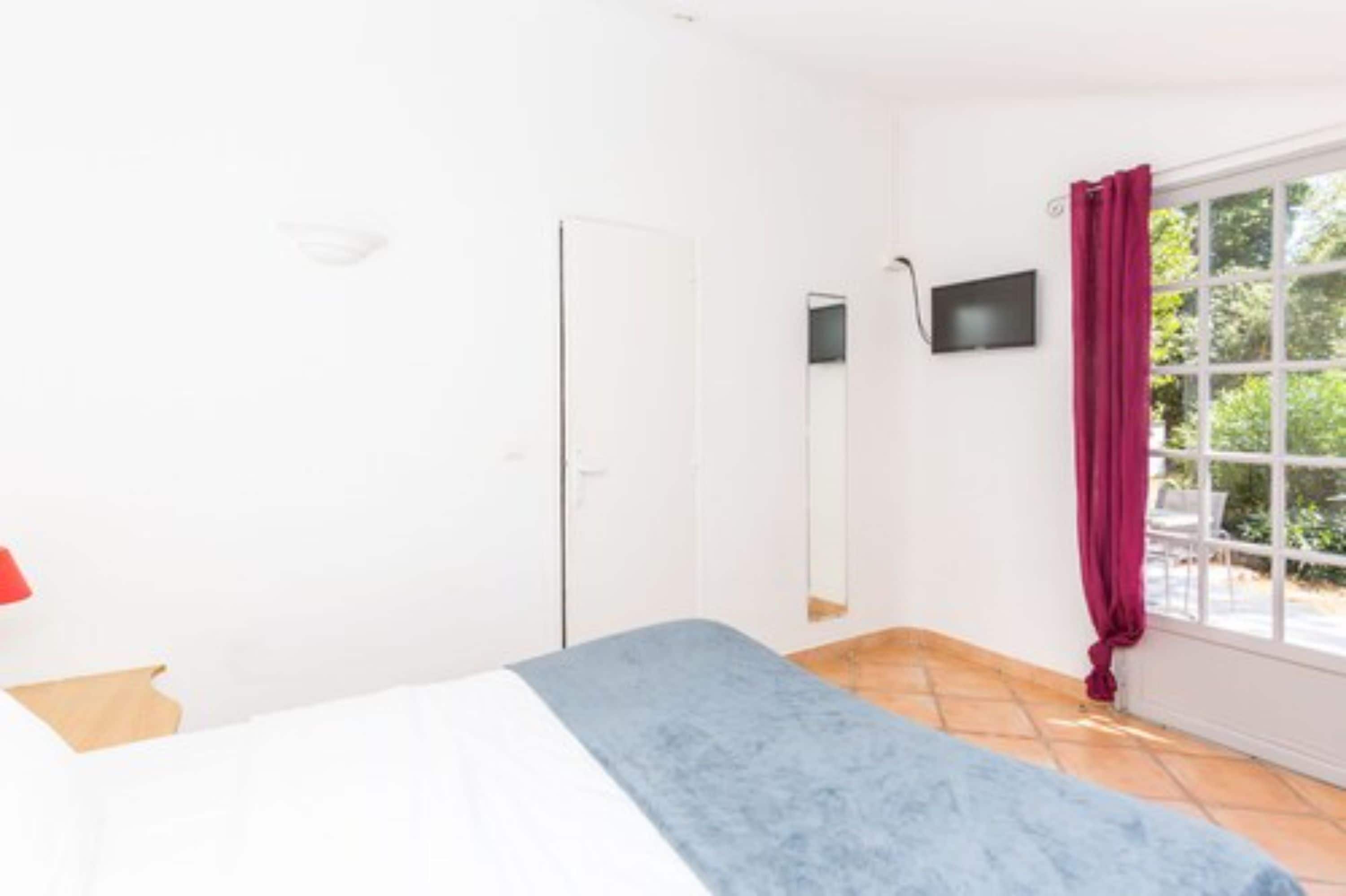 A bedroom with a bed, a window with a red curtain, a mirror, and a TV on the wall.