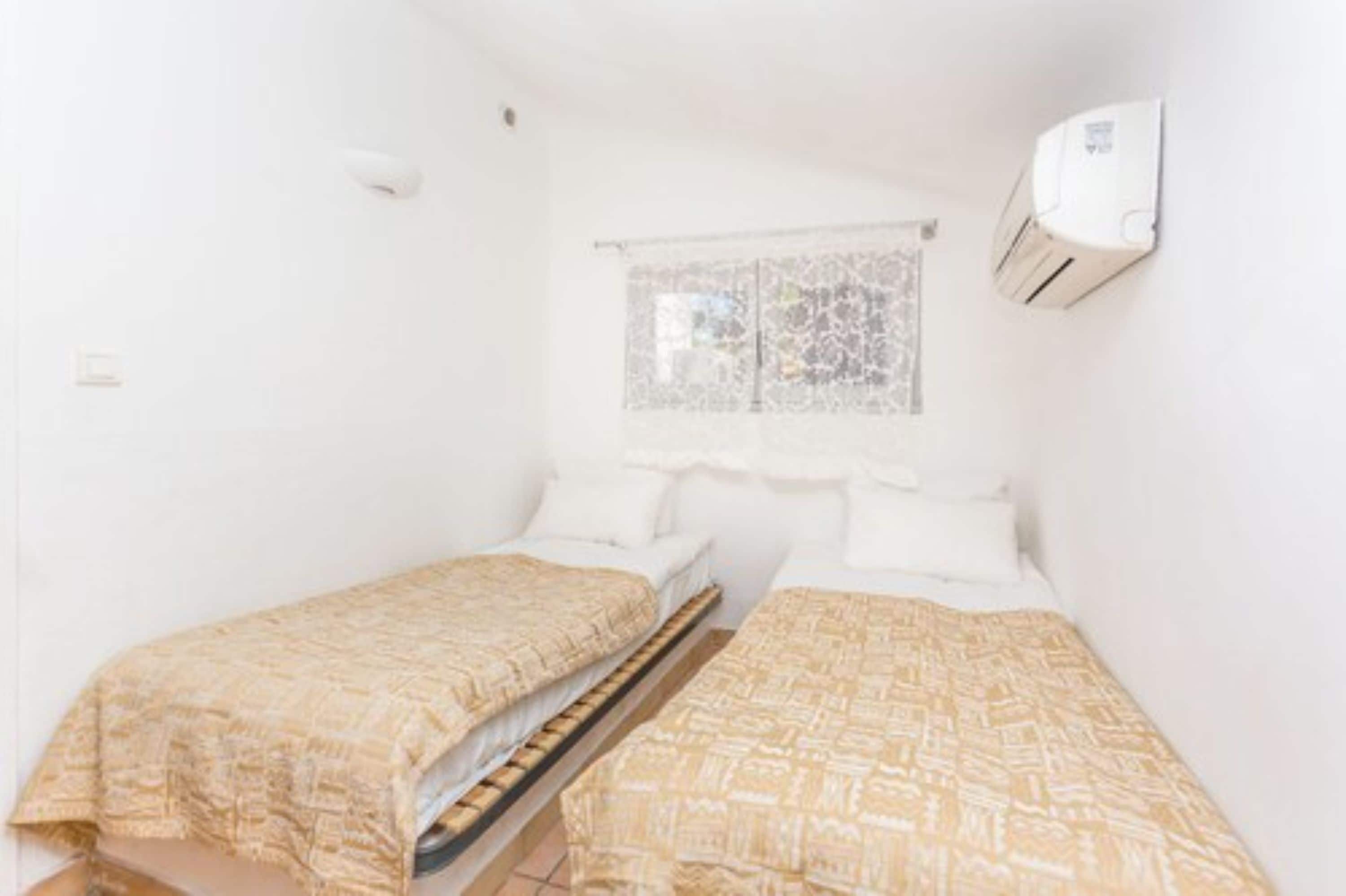 Una habitación pequeña y limpia con dos camas individuales, una ventana con celosía decorativa y un aire acondicionado instalado en la pared.
