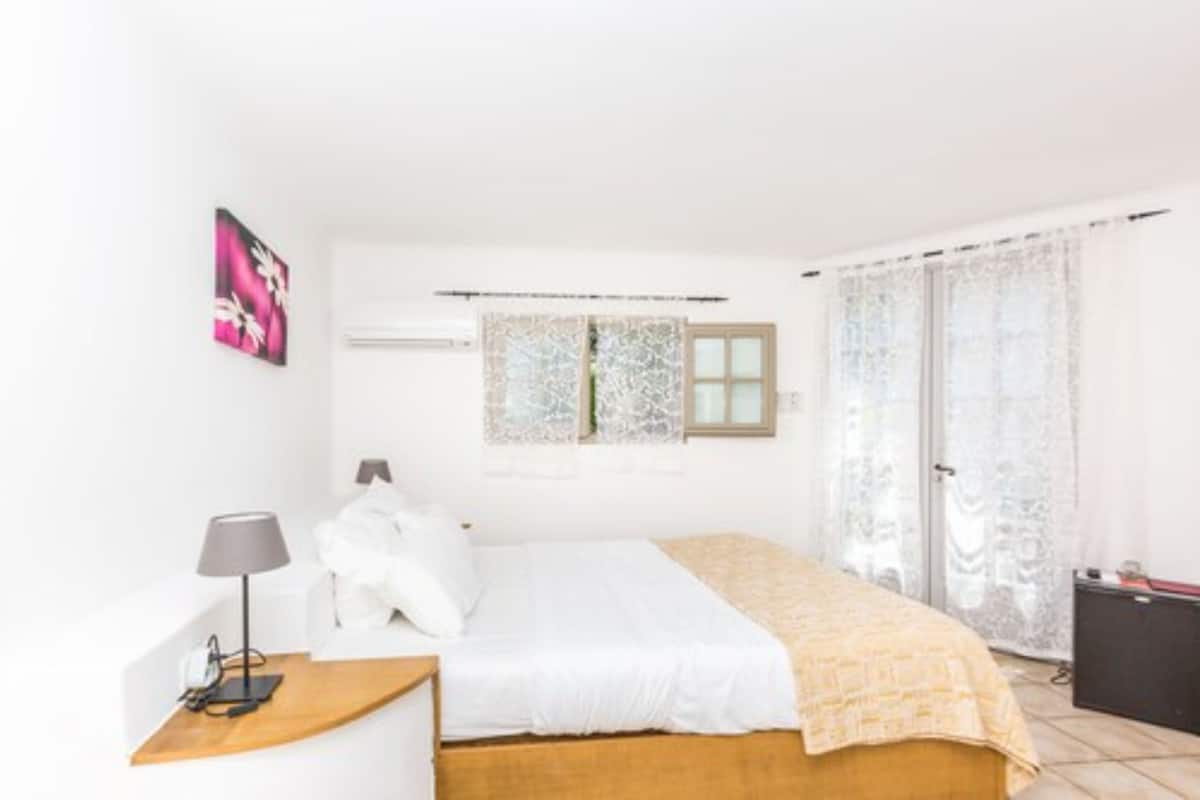 A bedroom with a bed, a bedside table, a lamp, a window with lace curtains, and a wall-mounted air conditioning unit.