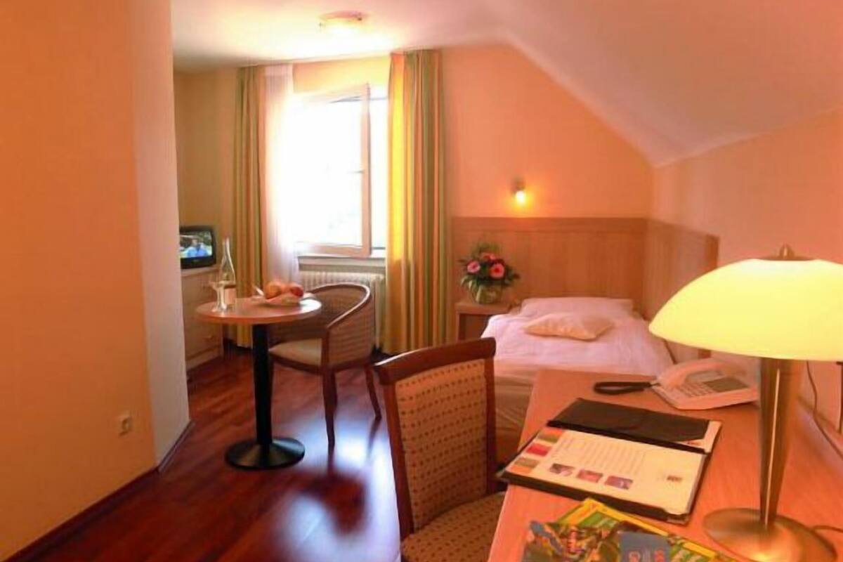 A hotel room with a bed, a desk with a lamp, a chair, a small table with a TV, and a window with curtains.