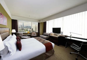 A hotel room with a bed, a desk, a chair, a TV, and a view of the cityscape.