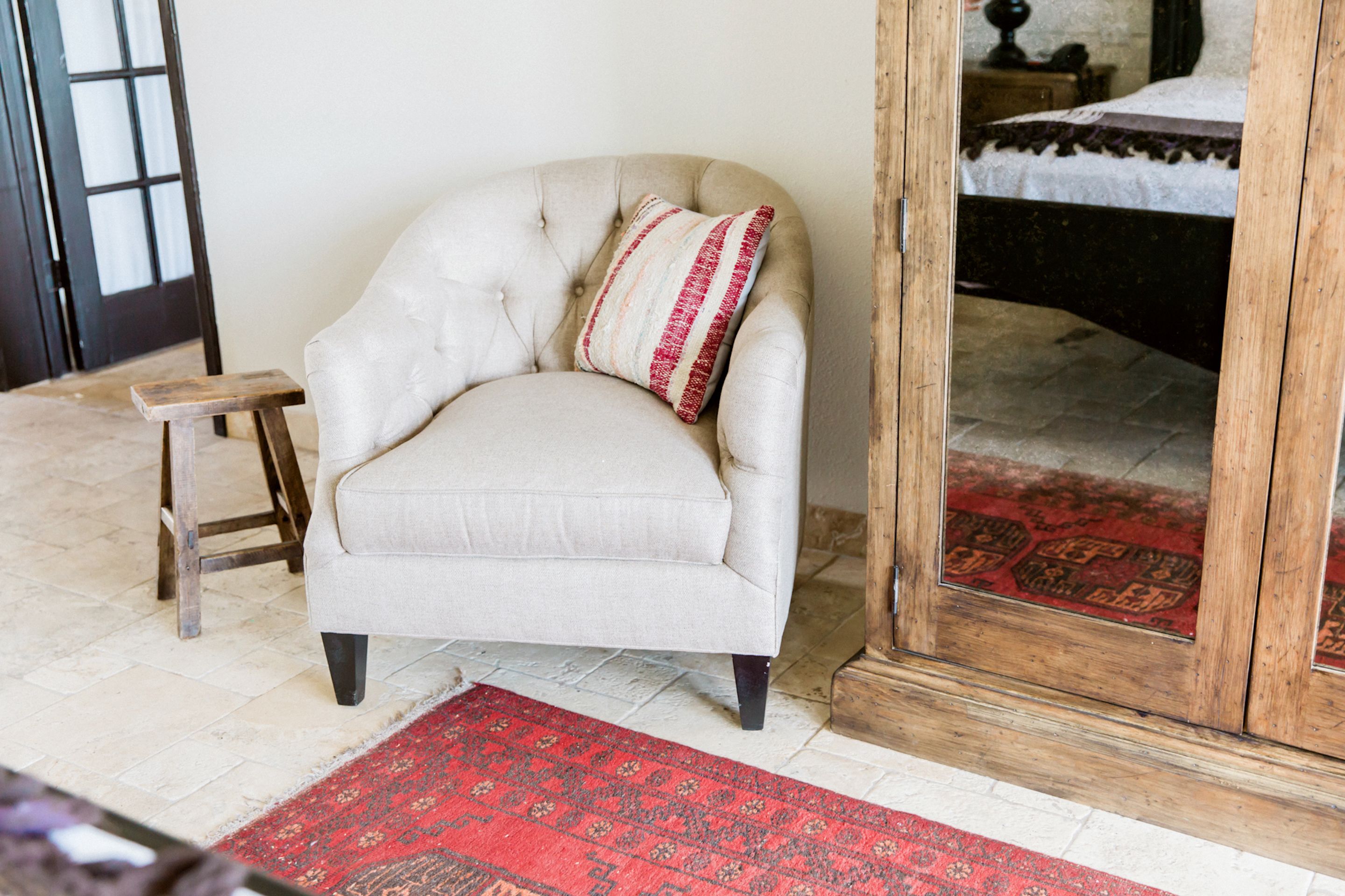 Une pièce comprenant un fauteuil capitonné, un petit tabouret en bois et un tapis rouge à motifs.